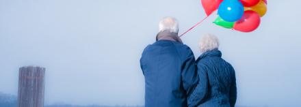 Photo of elderly couple holding red, yellow, green and blue balloons.
