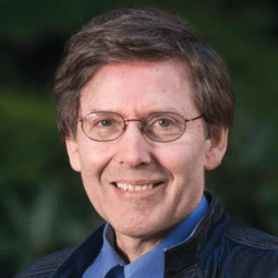 A man with glasses and short brown hair smiles at the camera. They are wearing a blue shirt and a dark jacket. The background is blurred greenery.