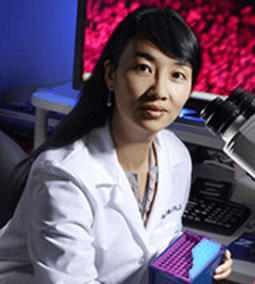 A woman in a lab coat holds a box with test tubes, sitting near a microscope in a laboratory setting.
