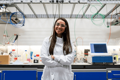 Smiling female scientist in a white lab coat standing in front of a laboratory bench with chemistry equipment. Courtesy Pixabay