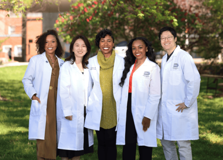 Five people wearing white lab coats stand side by side on the grass, representing a diverse team in a scientific setting.