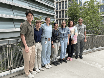 Seven people stand side by side on an outdoor balcony, smiling at the camera. They wear ID badges and casual clothes; glass-and-metal buildings and trees behind.