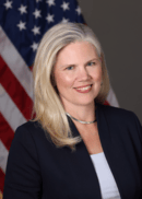 Woman with long blonde hair in a dark blazer, with a U.S. flag in the background.