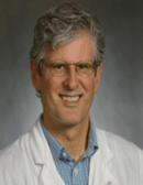 Man with gray curly hair and glasses wearing a white coat.