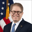 Man with round glasses in a suit and tie, with a U.S. flag in the background.