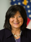 Woman with dark hair wearing a dark jacket and necklace, with a U.S. flag in the background.