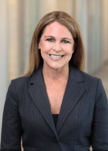 Professional portrait of a woman with long brown hair, wearing a dark blazer and smiling confidently in an office setting