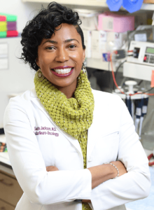 A woman in a lab coat with a green scarf stands in a laboratory, smiling.