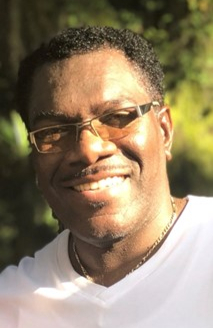A man smiling outdoors, wearing glasses and a white shirt, with sunlight illuminating his face and greenery in the background.