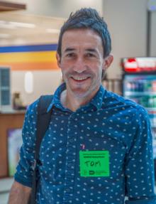 Man wearing a blue patterned shirt and green name tag smiling indoors.