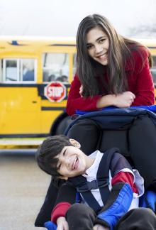 Picture of person with cerebral palsy sitting in a chair in front of a school bus along with a woman. Both people are smiling.