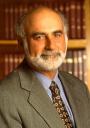 A headshot of a man with a beard, wearing a suit, framed in front of a bookshelf.
