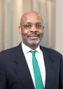 Professional portrait of a man with glasses and a short beard, wearing a dark suit, white shirt, and green tie, smiling in an office setting.