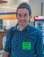 Man wearing a blue patterned shirt and green name tag smiling indoors.