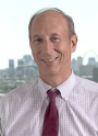 Smiling man in a dress shirt and tie, with a city skyline in the background.