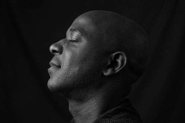 A man tilts his head back with his eyes closed against a dark background.