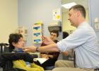Picture of patient examination. Patient in wheelchair is on the left. The examining doctor is on the right. In the middle of the picture is another doctor sitting and observing the examination.